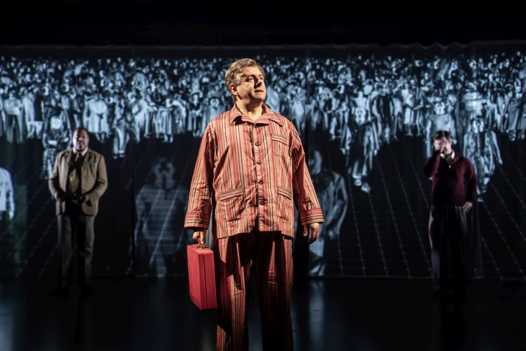 Michael Sheen stands center stage in striped pyjamas, holding a red briefcase, during a performance of Nye at the National Theatre. Behind him, a black-and-white projection of a marching crowd of doctors fills the backdrop.